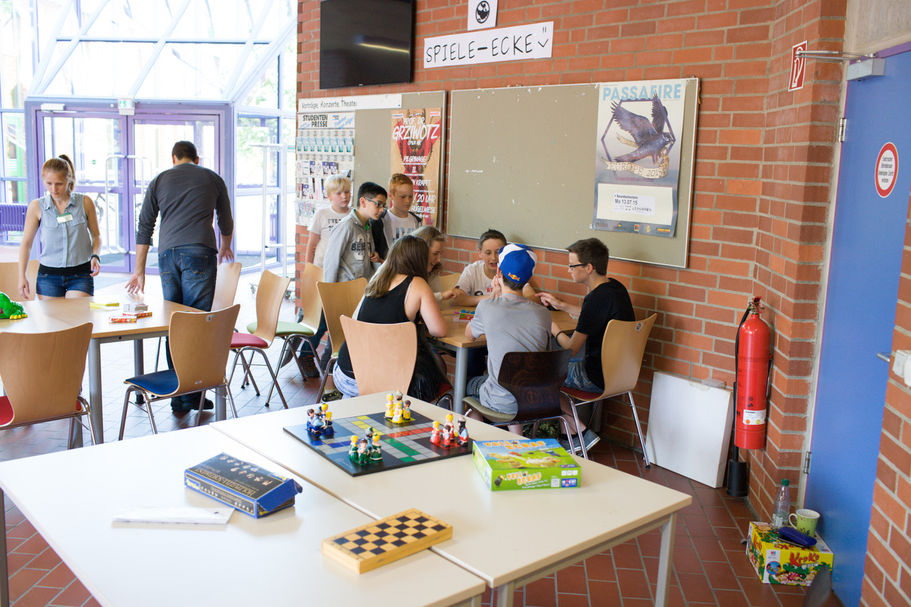 Tisch mit Spielen (Dame, Holzpuzzles, ...) im Foyer vor dem H 17 mit TdM-Teilnehmenden im Hintergrund